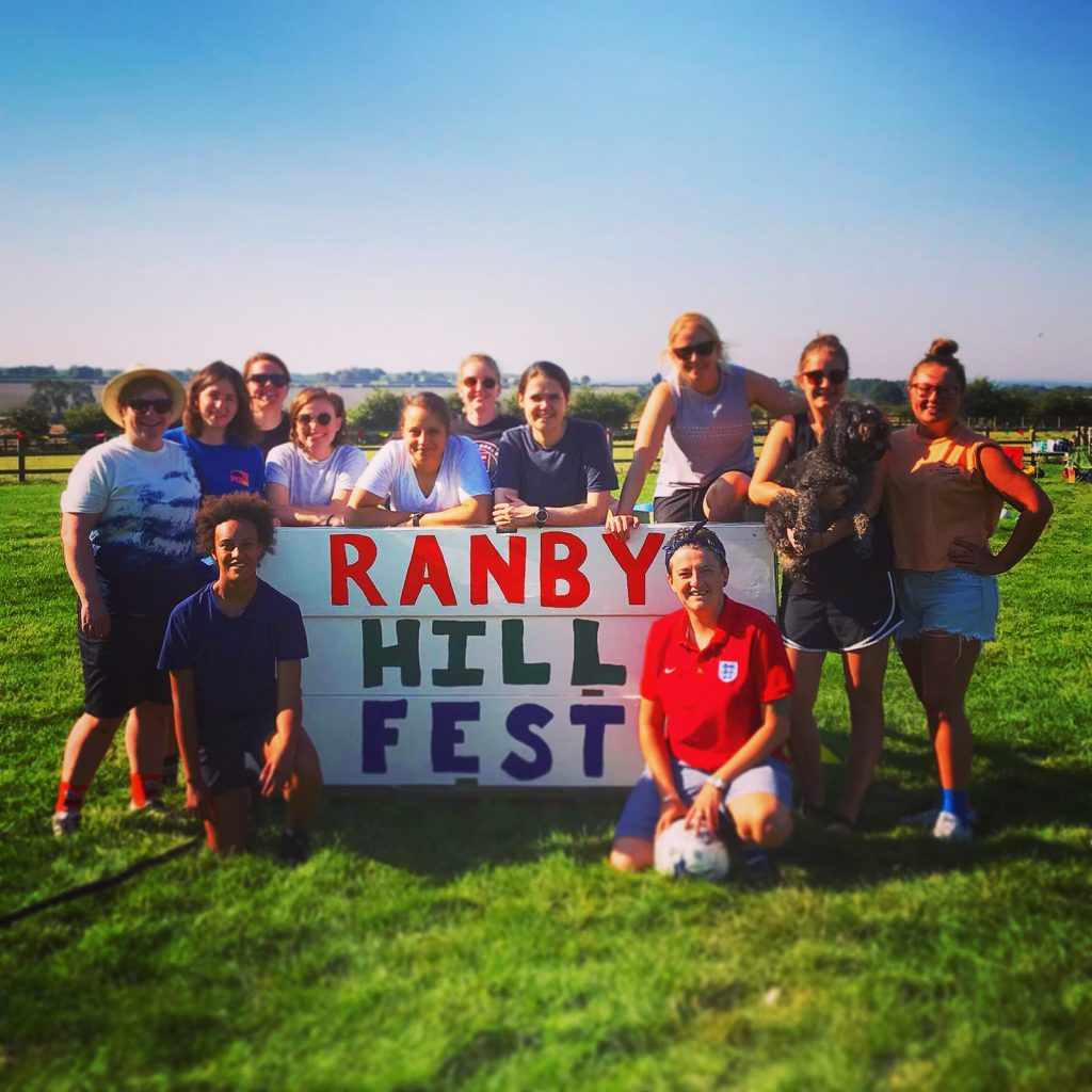 A photograph of the Ranby Hill Fest sign, with a group of 12 women standing around it in the open field. It is very sunny.