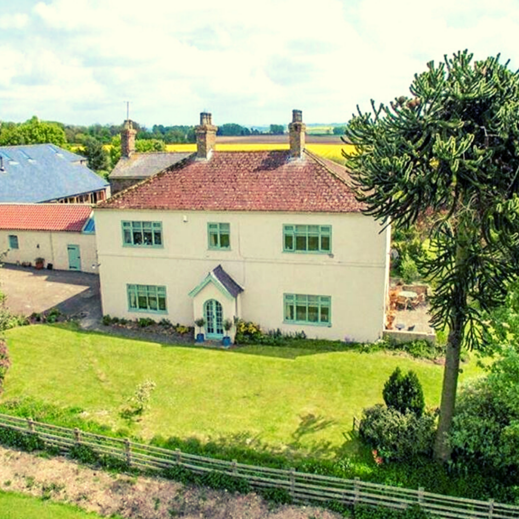 An ariel photograph of the farmhouse - a two storey building with two chimneys painting in white with green window trims. You can see the gardens and patio surrounding the house.